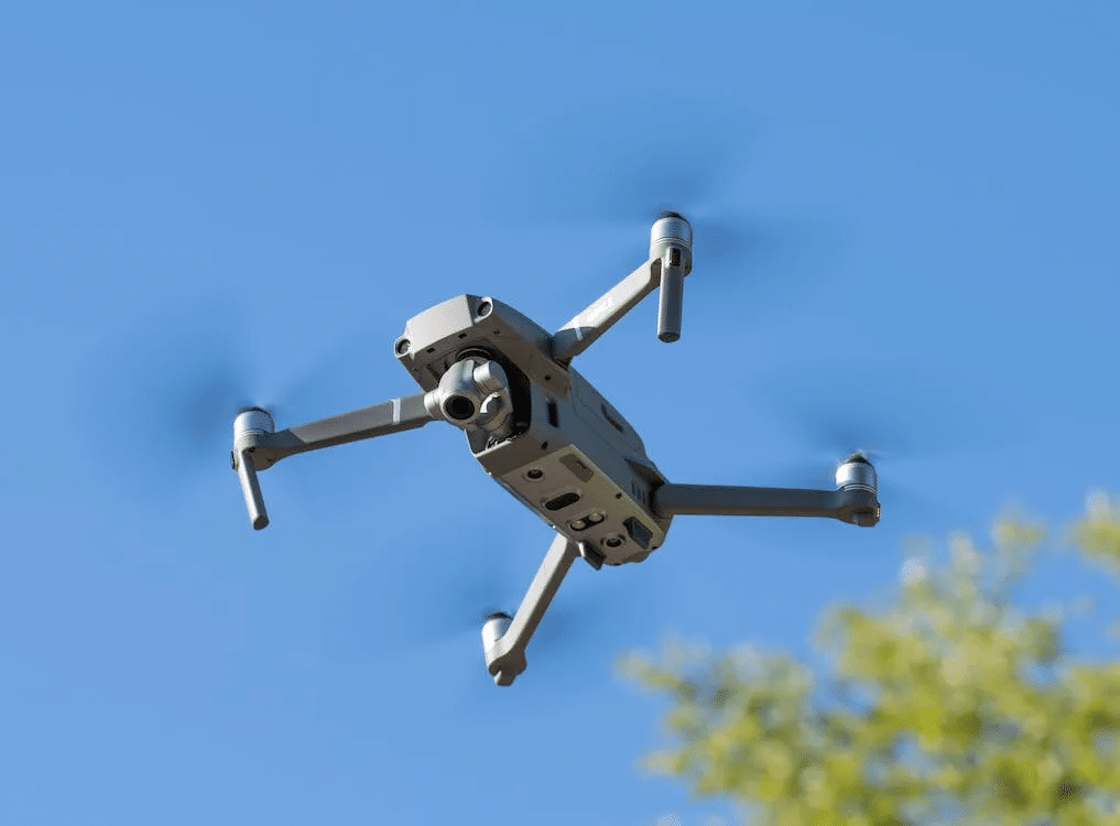 Gray drone flying in a blue sky. Drone repair may be needed if there's a crash. ER4Gadgets